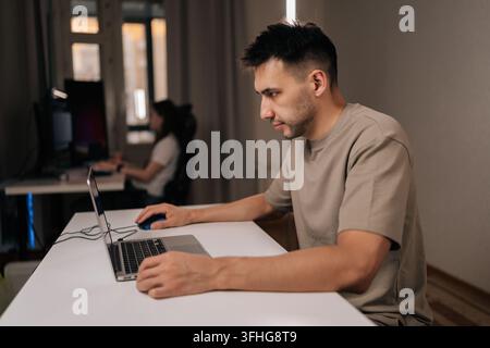 Side view of software developer looking on laptop screen, using mouse and typing keyboard while working from modern desk, with female colleague in Stock Photo