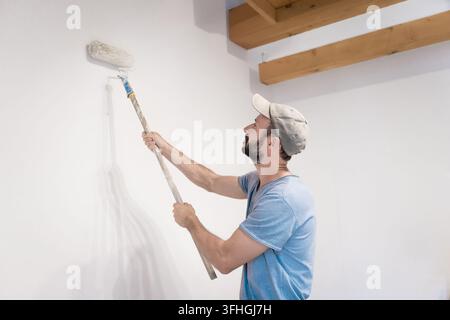 young man focused while painting interior wall with roller brush smiling, millennial doing home renovation, diy home improvement, home decor, interior Stock Photo