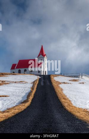 View of a white church with a vibrant red roof situated on a snowy hill at the end of a gravel road under a dramatic cloudy sky in Iceland. Stock Photo