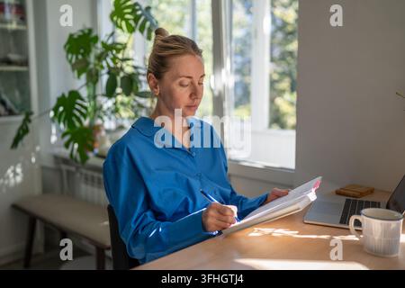Concentrated positive woman freelancer planning day and tasks while sitting at table in kitchen. Stock Photo