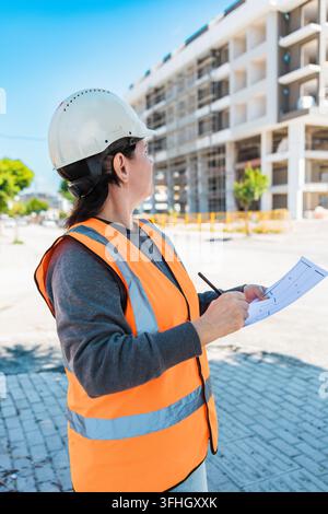 A young Hispanic woman in a safety vest and hard hat inspects a construction site. She holds a clipboard and looks at the building under construction. Stock Photo