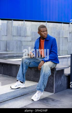 A young Black man sits on concrete steps, wearing a blue jacket and orange shirt. He holds a smartphone and looks thoughtfully into the distance. Stock Photo