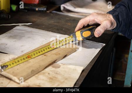 The senior craftsman uses a yellow tape measure to verify dimensions of lumber in a dusty workshop, symbolizing skilled manual labor, home improvement Stock Photo