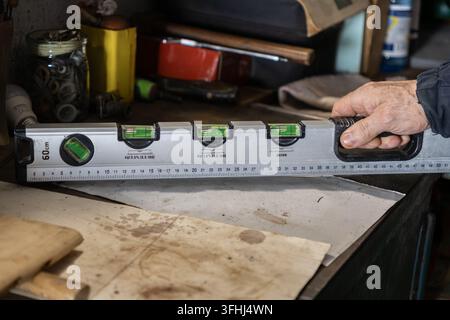 An experienced worker uses a long metal spirit level to measure a flat surface in a cluttered garage, representing home renovation, DIY improvement, a Stock Photo