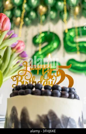 A festive cake featuring a golden twenty-fifth birthday topper with a lit candle, set against a soft bokeh backdrop of balloons and flowers for a joyo Stock Photo