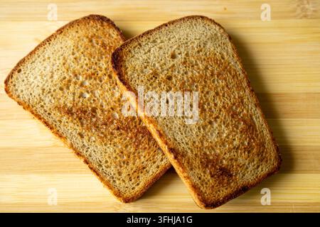 Two warm toasted slices of whole grain bread lie together on a smooth light wood surface. The close up detail highlights the golden brown texture. Stock Photo