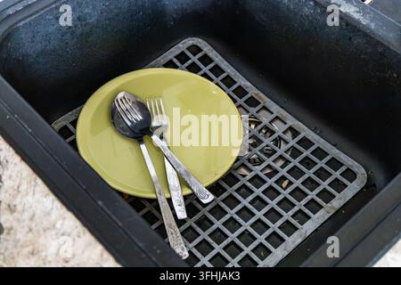 Yellow ceramic plate and stainless steel utensils rest inside a dark kitchen sink. A rubber sink mat supports the unwashed dishes under bright natural Stock Photo