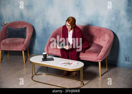 Accountant woman on pink sofa calculating budgets with tablet and receipts spread on table, focused analytical mood Stock Photo