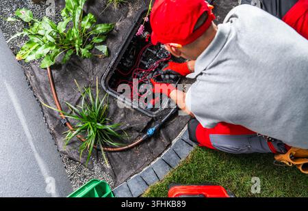 Overhead view of a technician working on valves and wiring inside an open irrigation valve box beside a lawn and planted border. Stock Photo