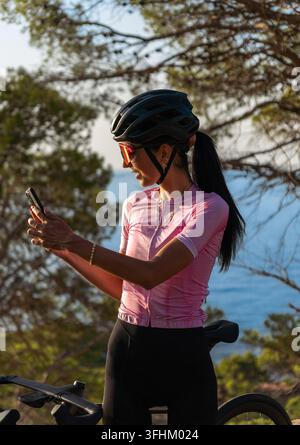 A young female cyclist using her smartphone for a video call and laughing happily, standing next to her road bike on a coastal road near Tossa de Mar Stock Photo