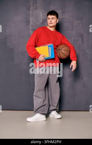 A fashionable young man stands confidently while holding a basketball and colorful notebooks. Stock Photo