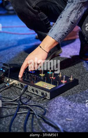 Musician hand adjusts illuminated knobs on guitar effects pedalboard beside coiled cables across blue stage floor, showing detailed live performance e Stock Photo