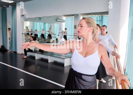 Adult female ballerina with blonde hair and focused expression extends arm beside barre in bright mirrored studio, with adult dancers rehearsing benea Stock Photo