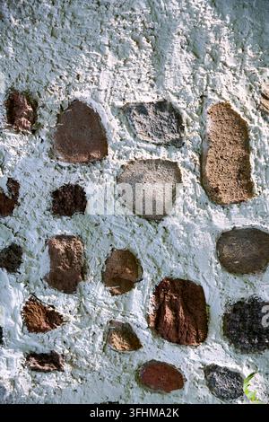 Rough stone wall covered with pale blue plaster holds uneven brown and gray rocks beneath soft sunlight, creating tactile architectural texture for ba Stock Photo