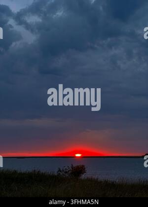 Dramatic red sunset glowing across the horizon beneath dark storm clouds, with calm water and natural grass in the foreground. Atmospheric landscape s Stock Photo