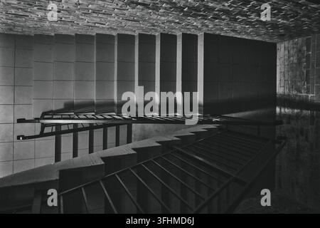 Geometric staircase descends through concrete interior beneath textured ceilings and angular railings, with stark monochrome light creating architectu Stock Photo