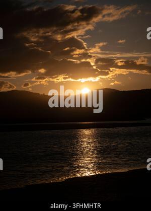 Golden sun setting behind mountains creating a warm glow and reflections on the water Stock Photo