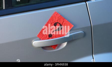 A crumpled red paper note with a black question mark printed on it tucked behind the silver door handle of a gray car. Stock Photo