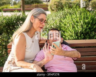 Mother and adult son with disability using a smartphone on a park bench Stock Photo