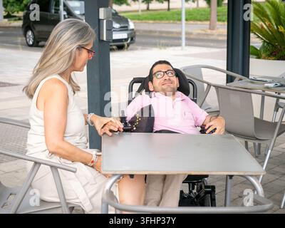 Disabled man sitting in electric wheelchair at outdoor cafe with his caregiver Stock Photo