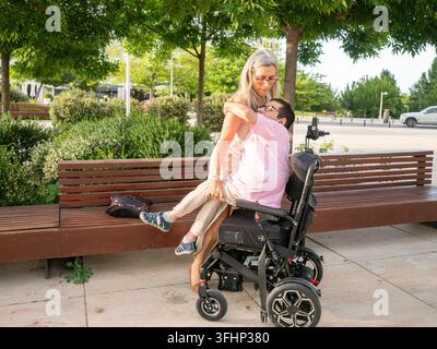 Woman helping disabled man transfer from his electric wheelchair in a park setting Stock Photo