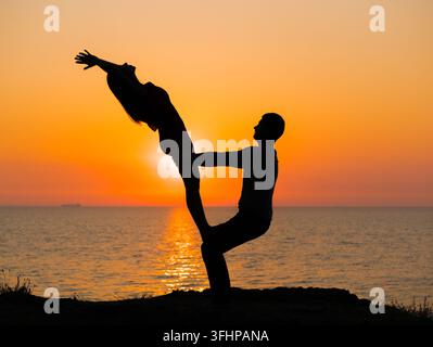 Silhouette of young woman and man doing pair yoga on sea beach at sunset. Meditation. Couple practicing acro yoga. Flexibility w Stock Photo