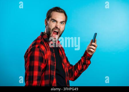 Angry modern hipster in red plaid shirt screaming down his mobile phone. Stressed and depressed man on blue background. Stock Photo