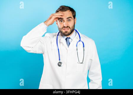 Portrait of young puzzled man in professional medical white coat isolated on blue studio background. Forgetful doctor with beard and stethoscope. He h Stock Photo