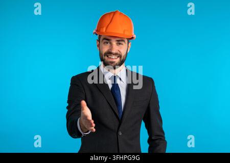 Young engineer businessman in construction helmet holds out his hand for greeting, handshake. Man with beard in corporative suit over blue background Stock Photo
