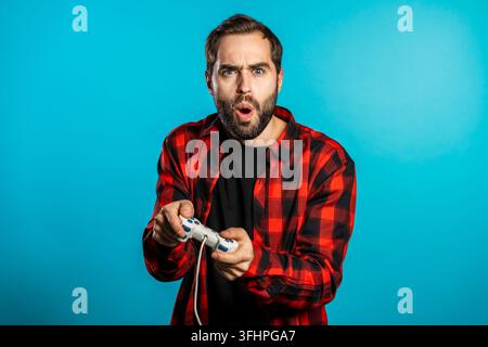Handsome young man playing video exciting game on Tv with joystick on blue studio wall. Using modern technology Stock Photo