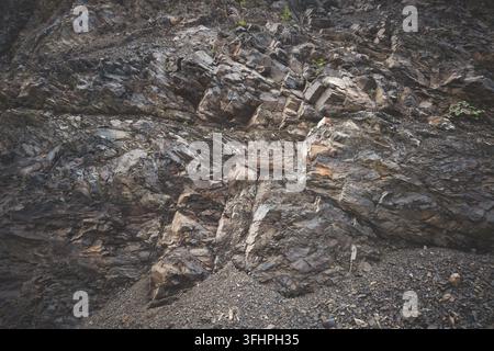 A close-up texture of a rugged cliff with distinct layered sedimentary rock with sharp, jagged edges and deep cracks. Abstract dark background Stock Photo