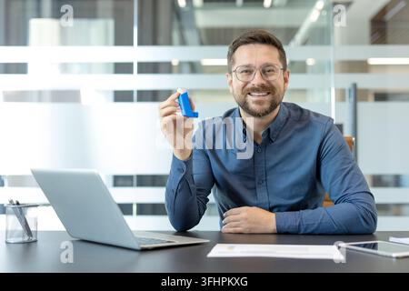 Asthma management and respiratory health, smiling man holding an inhaler at his office desk with a laptop for workplace wellness and medical care. Stock Photo
