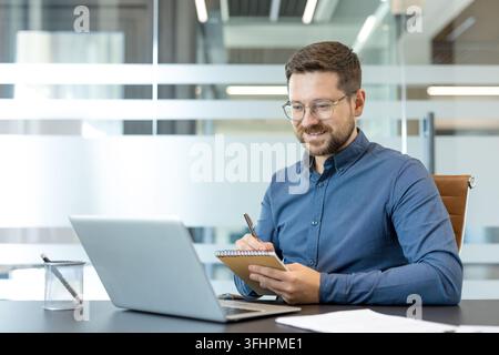 Professional businessman working on laptop and taking notes in modern office, remote work and corporate productivity concept. Focused male employee using computer and writing in notebook for business. Stock Photo