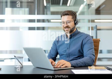 Customer support and remote work, smiling man with headset using laptop for video call in modern office. Professional communication and digital connectivity. Stock Photo