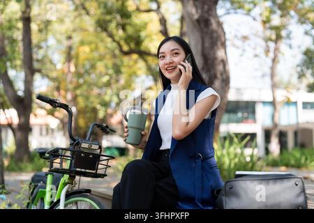 Asian businesswoman with bicycle using smartphone and sitting outside the office building. Woman commuting on bike go to work. Eco friendly vehicle, s Stock Photo