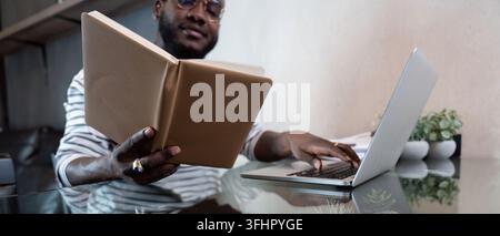 Young african man using laptop at home, black male looking at read book relaxing on leisure with work sit on glass table in living room. Stock Photo