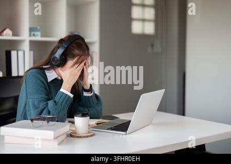 Young woman feeling stressed while wearing headphones and using a laptop in a home office Stock Photo
