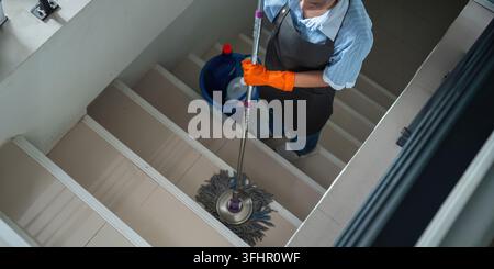 A cleaning woman is standing inside a building holding with a blue tank on the side chemicals and facilities for tidying up in her hand. Stock Photo