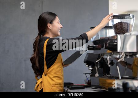 Smiling barista in yellow apron using coffee grinder in a modern cafe, holding a digital tablet. Stock Photo