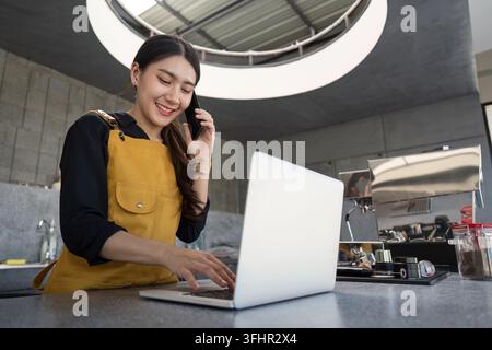 Young Woman Barista Working on Laptop and Talking on Phone in Modern Coffee Shop Stock Photo