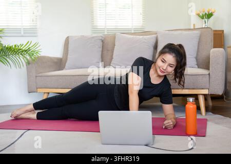 Woman Exercising at Home with Laptop, Doing Side Plank on Yoga Mat in Living Room Stock Photo