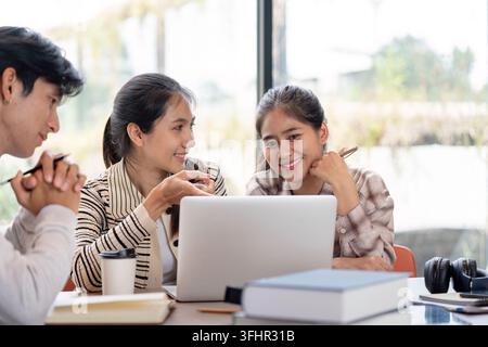 Three students working together on a laptop, sharing ideas and studying in a bright, modern environment with books and coffee. Stock Photo