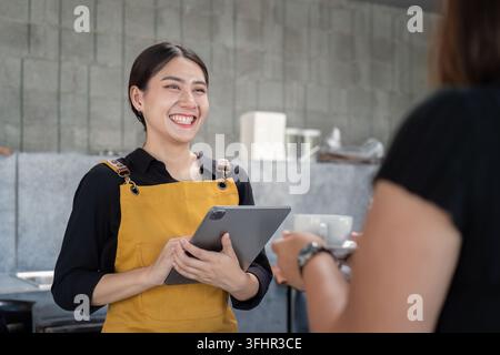 Friendly Barista in Yellow Apron Smiling and Taking Order on Tablet in Modern Coffee Shop Stock Photo
