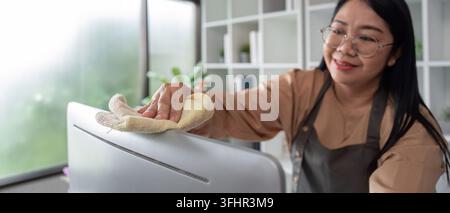 Professional Housekeeper Cleaning a Modern Home Office with a Smile, Wearing Apron and Glasses, Using a Cloth to Wipe Down Surfa Stock Photo