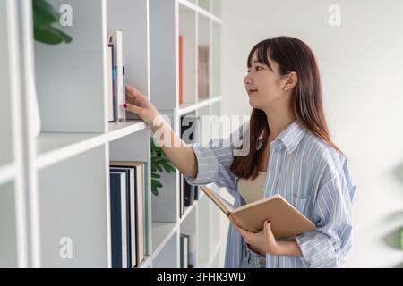 Young Student Studying Online at Home, Reaching for a Book on a Shelf, Embracing Modern Education Technology and Remote Learning Stock Photo