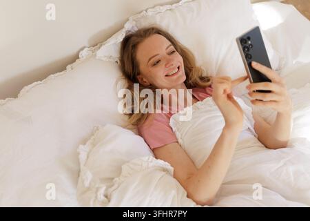 Smiling woman uses a smartphone while relaxing in bed, representing bedtime AI assistance, personal technology, online conversation, and pre-sleep com Stock Photo