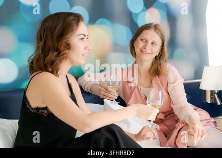 Two women are sitting on a bed and drinking wine Stock Photo