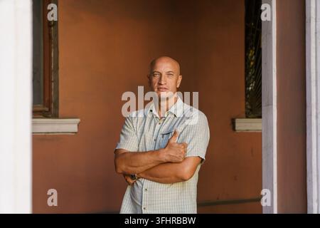 A balding middle-aged man in a plaid shirt stands against a painted wall Stock Photo
