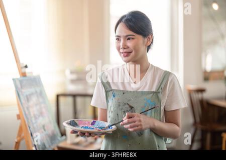 Smiling female artist holding a palette and paintbrush, ready to create in a serene art studio Stock Photo