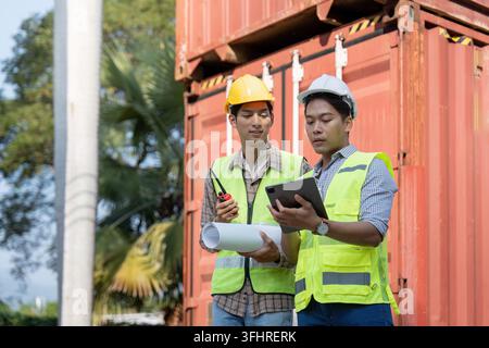 Two engineers in safety vests and helmets examine logistics plans on a tablet and blueprints while standing near shipping containers, emphasizing coll Stock Photo